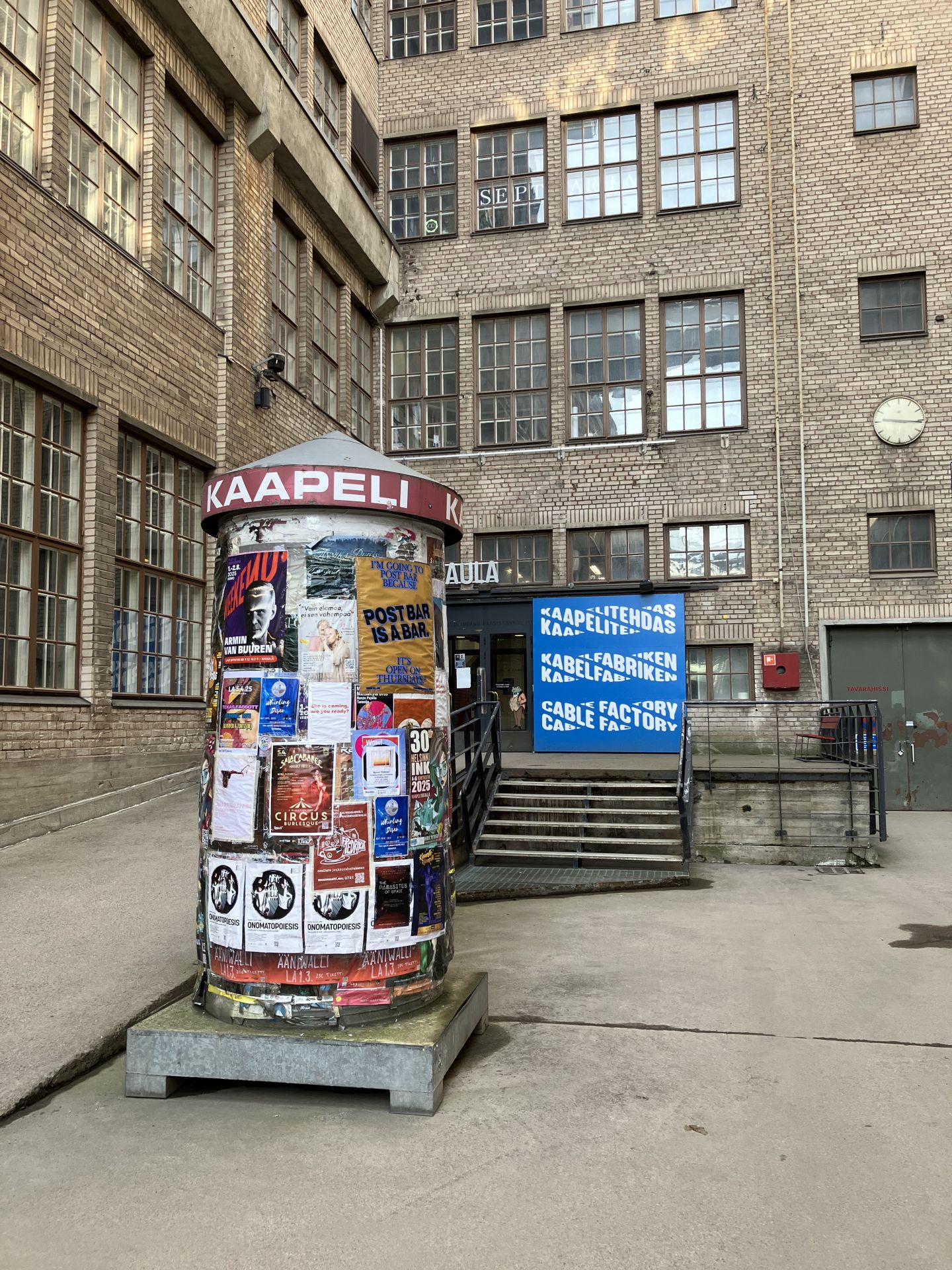 En närbild tagen på innergården på ett tegelhus med många fönster. Framför huset finns en rund reklampelare med många olika afficher på och texten Kaapeli..