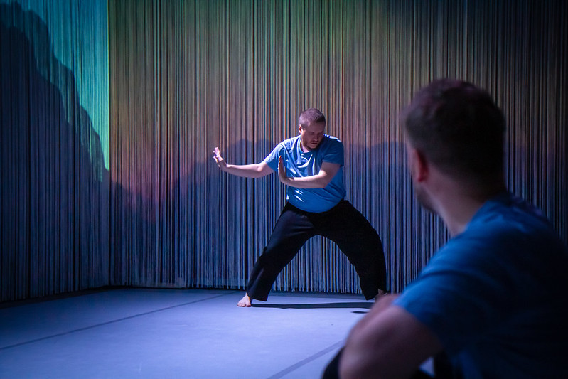 DuvTeatern dancer Emil Nordman dances with his arms dramatically extended to one side. Nordman wears a light blue t-shirt and loose black pants. Dancer Eero Vesterinen sits in front, observing Nordman.