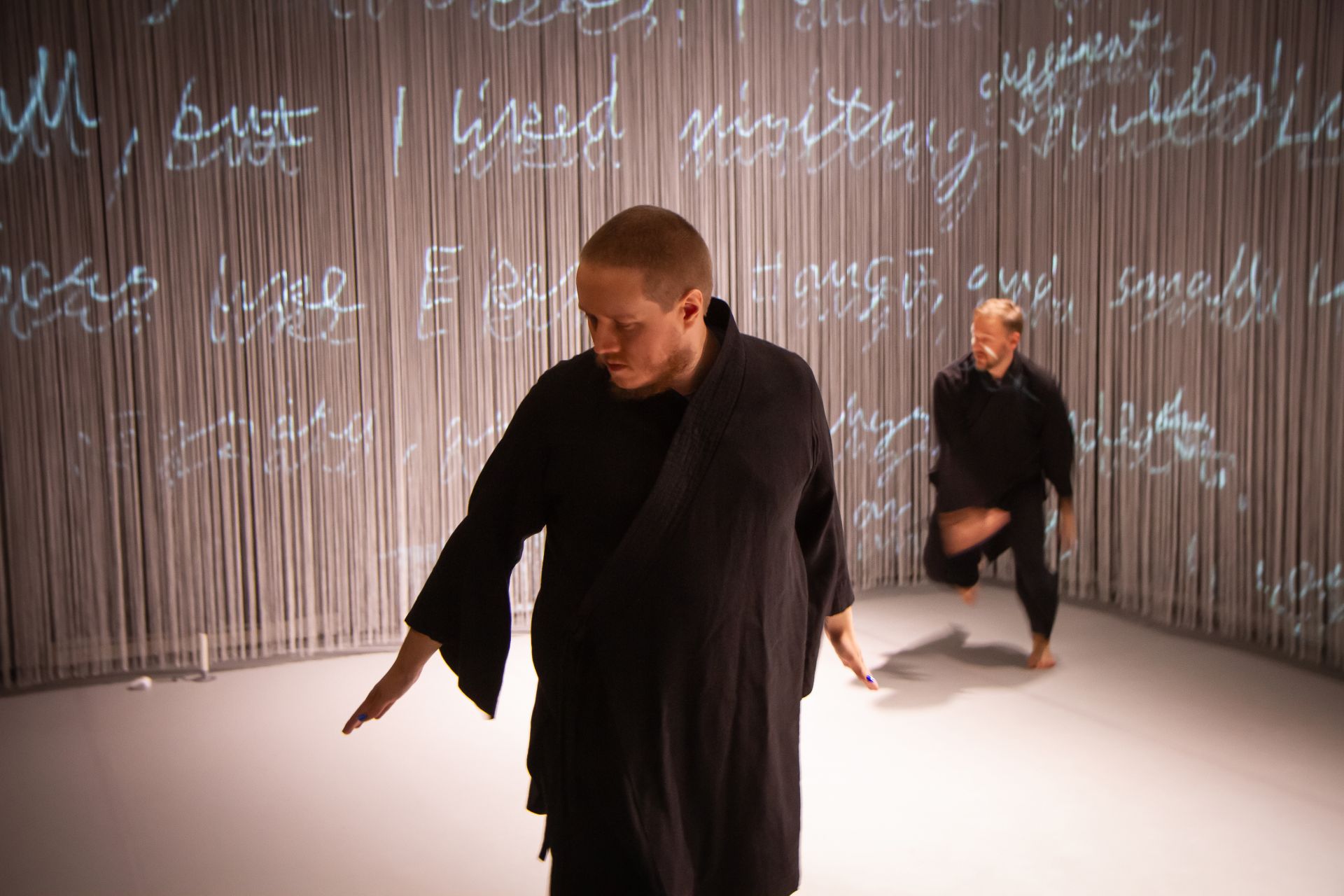 DuvTeatern dancer Emil Nordman stands in the foreground wearing a black kimono, dancing and looking down at the ground. In the background, dancer Eero Vesterinen dances in his own black kimono. The backdrop reflects Emil's diary.