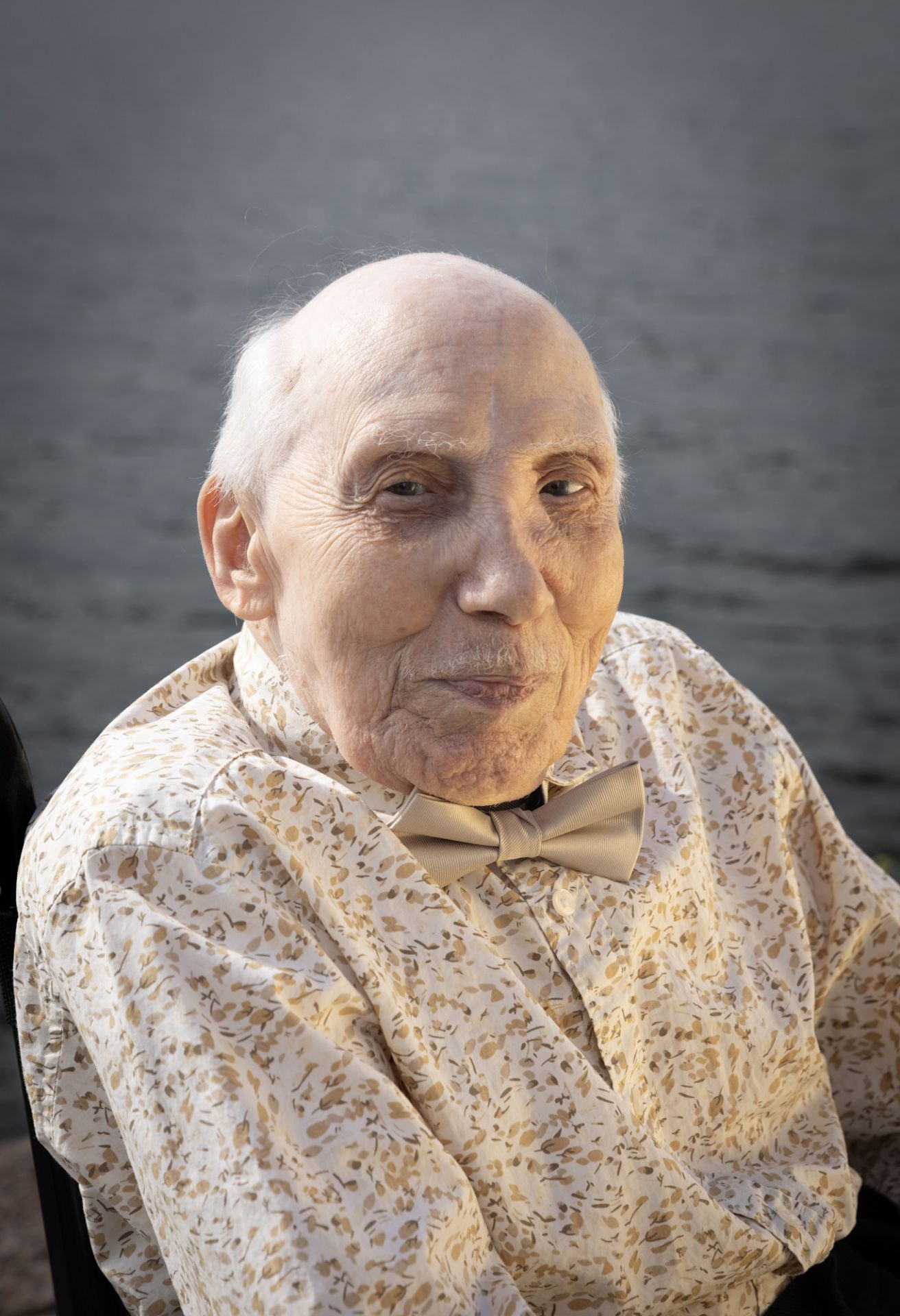 Till vänster ett porträtt av en äldre man, iklädd skjorta och fluga, som tittar rakt in i kameran. I bakgrunden hav.