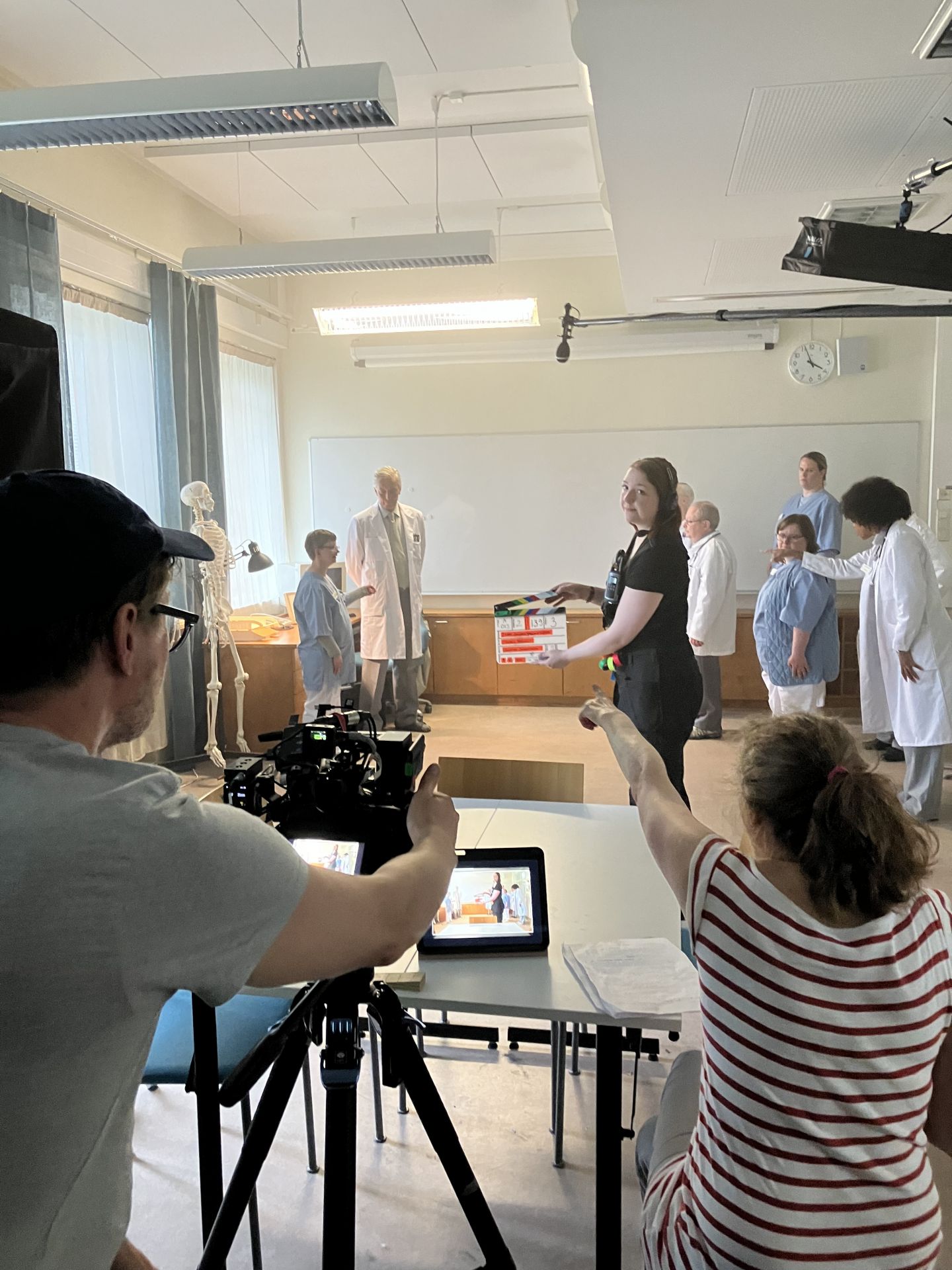 Bild från filminspelning. Skådespelare och filmteam förbereder en scen, medan en kameraman filmar en grupp personer i vita läkarrockar och andra scenkläder. En person håller upp en filmklappa framför kameran.